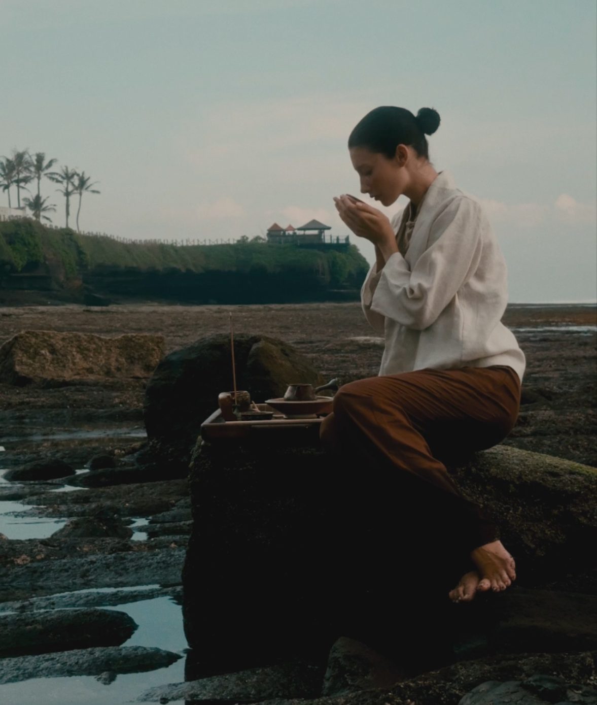 A woman sitting on rocks by the sea, sipping from a cup